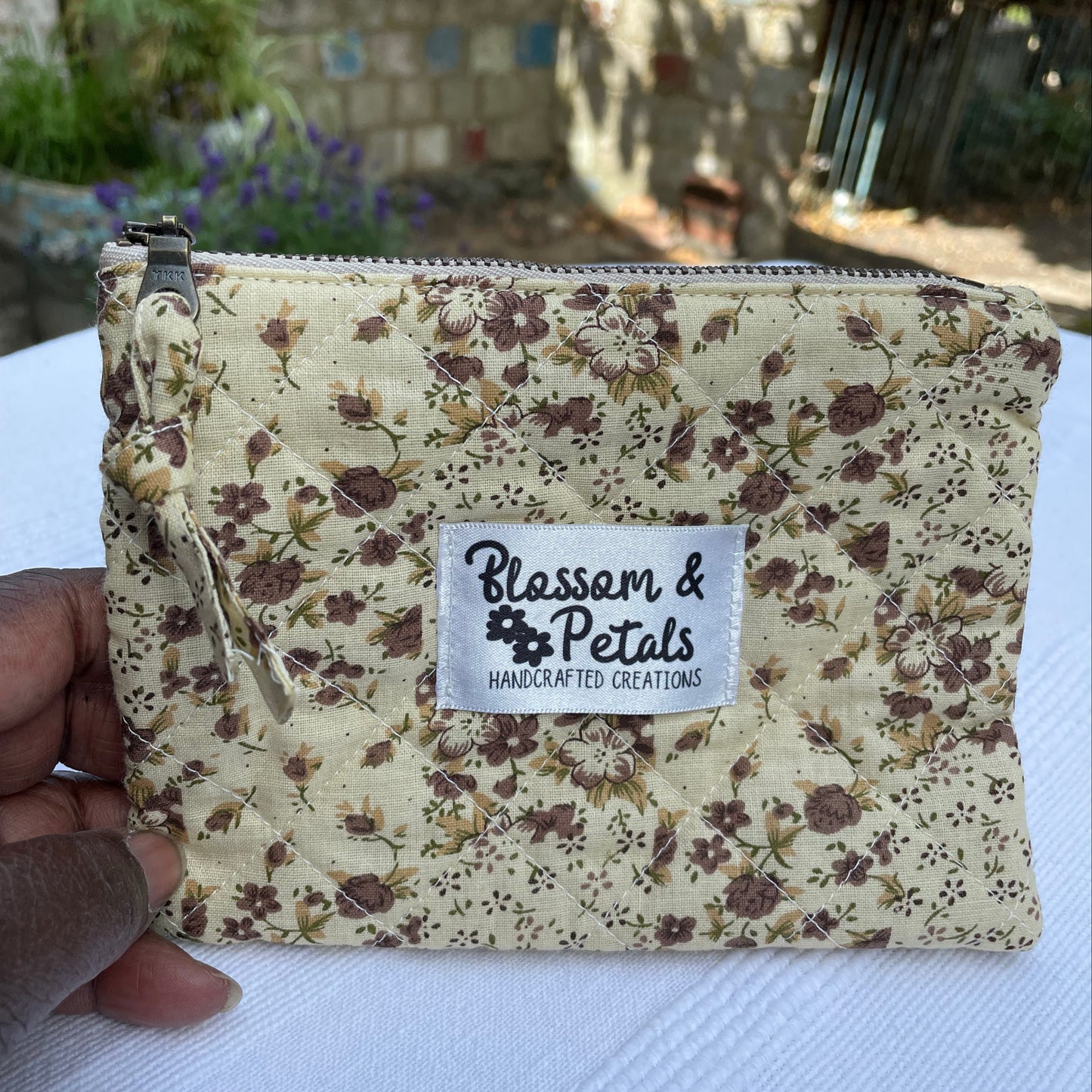 Beige Floral-patterned pouch with a visible brand label held by a hand on a textured surface.
