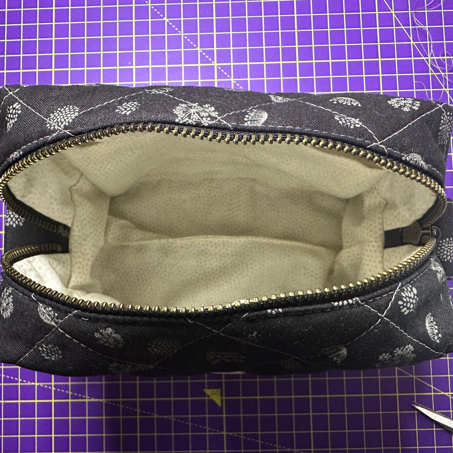 Black quilted makeup bag /pouch with grey floral pattern on a purple cutting mat