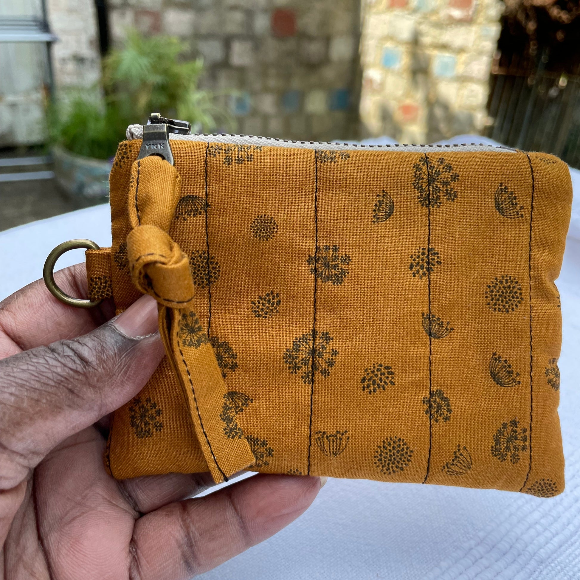 Hand holding a caramel coloured fabric pouch with floral patterns against a blurred outdoor background.
