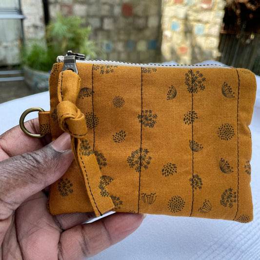 Hand holding a caramel coloured fabric pouch with floral patterns against a blurred outdoor background.