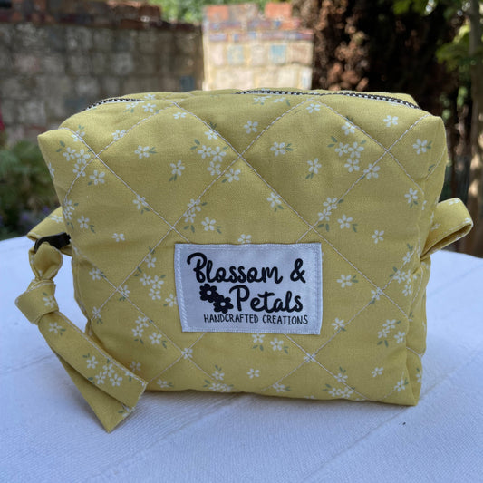 Yellow quilted pouch with white floral pattern and "Blossom & Petals Handcrafted Creations" label, styled on a white background outdoors