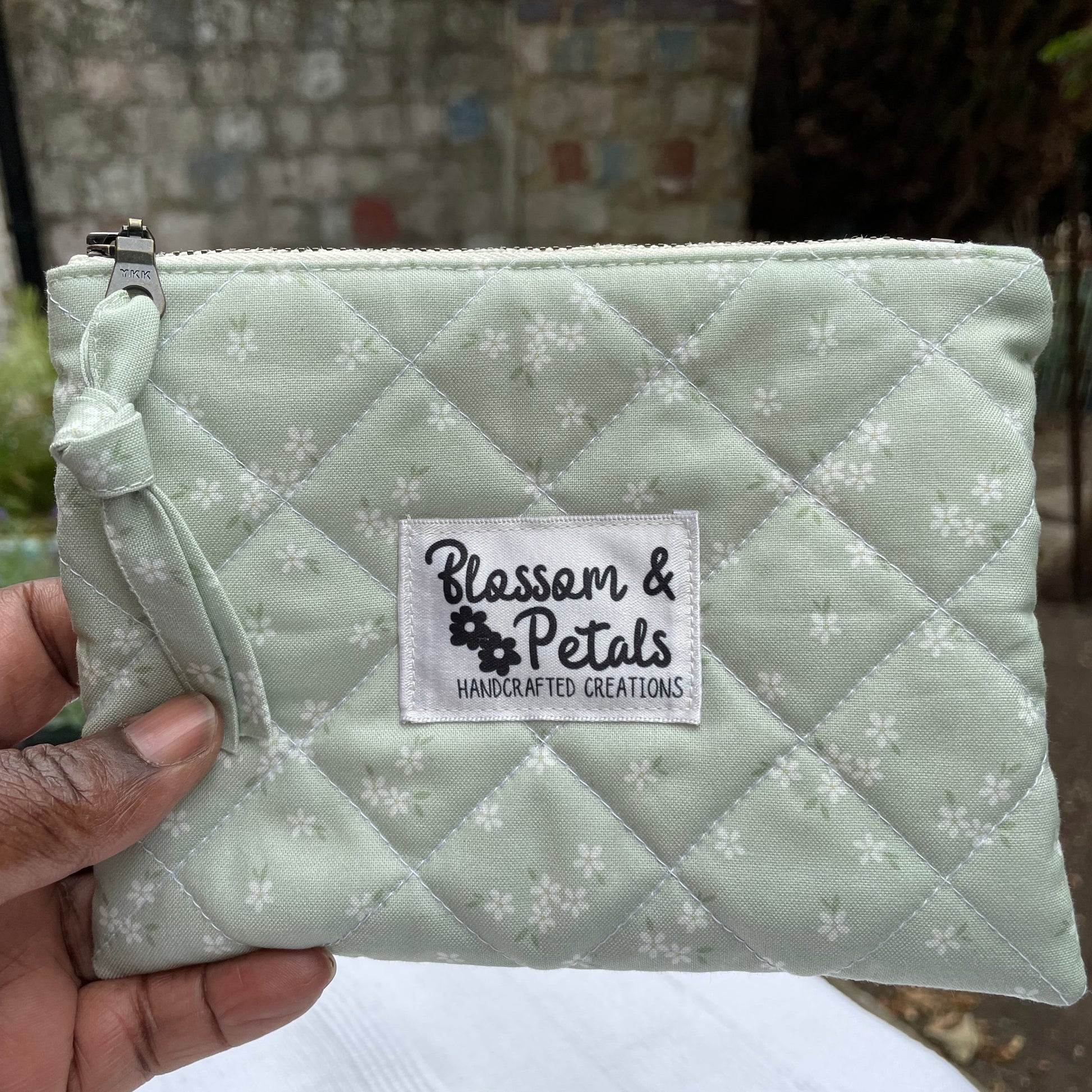 Quilted sage green pouch with floral pattern held by a hand, brand label visible.