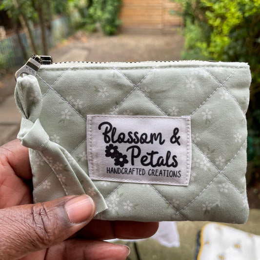 Quilted sage green pouch with a visible brand label held by a hand outdoors