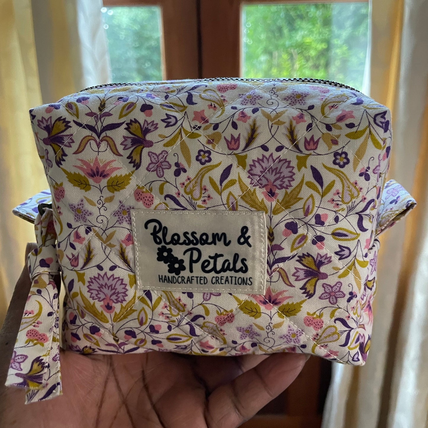 Handmade quilted pouch with cream background and purple floral print, displayed in hand with Blossom & Petals branding
