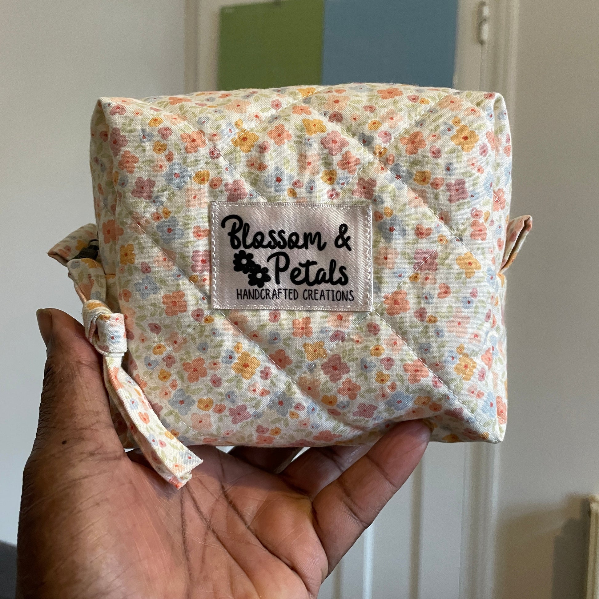 Hand holding a pastel floral quilted zipper pouch with soft peach, yellow, and blue flowers and Blossom & Petals handmade label.