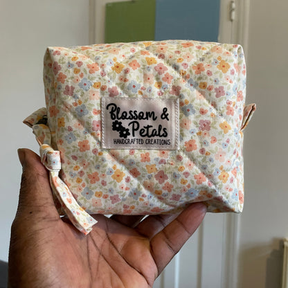 Hand holding a pastel floral quilted zipper pouch with soft peach, yellow, and blue flowers and Blossom & Petals handmade label.