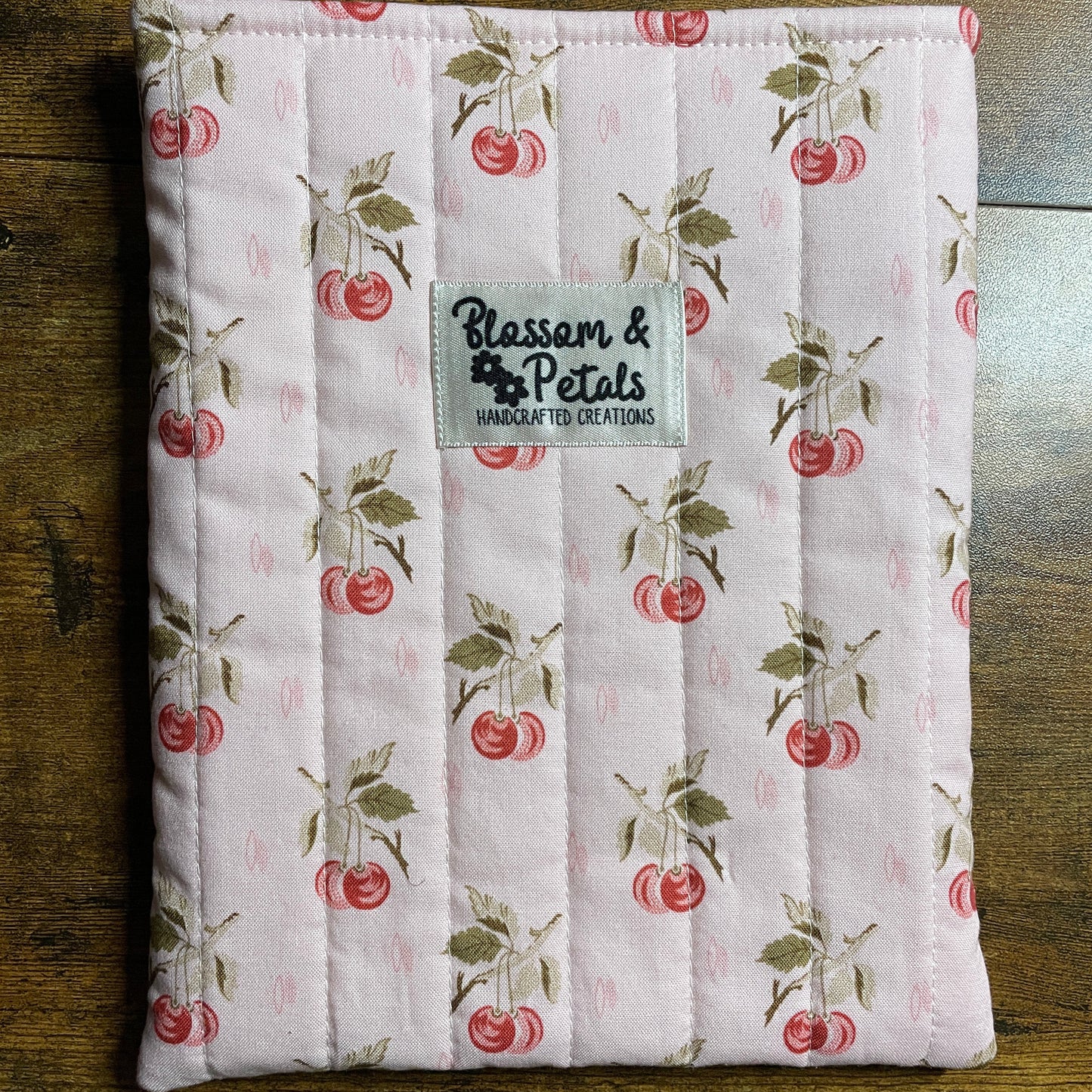 Handmade quilted pink book sleeve with cherry print fabric and Blossom & Petals label, padded fabric book cover for protecting books and journals.