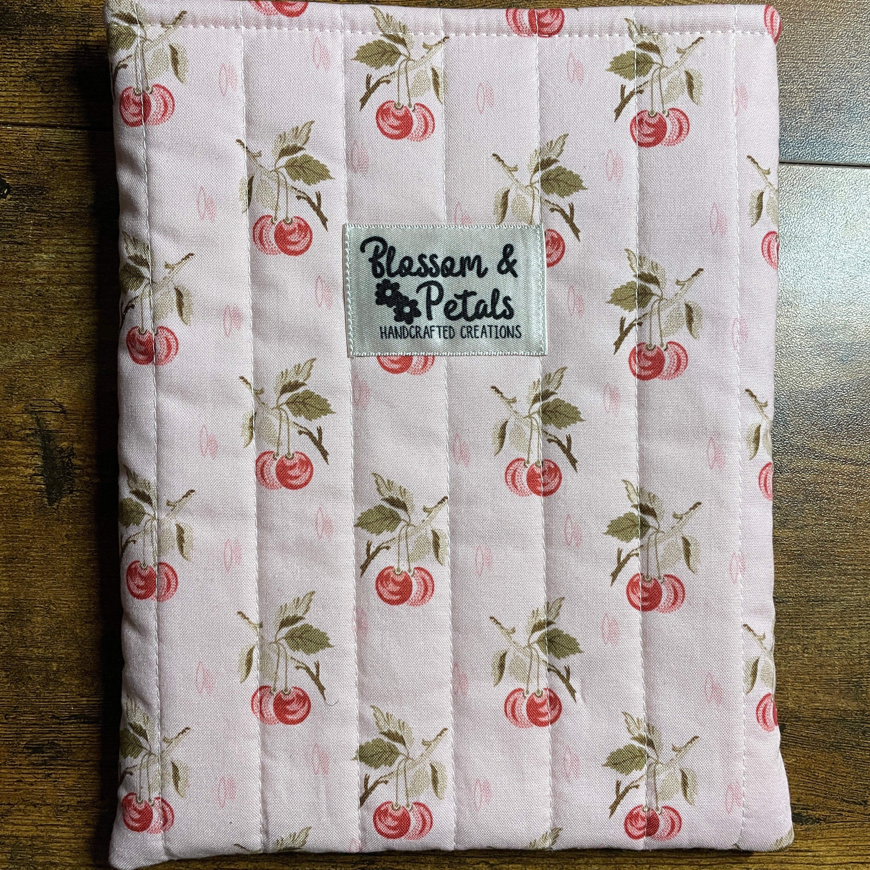 Handmade quilted pink book sleeve with cherry print fabric and Blossom & Petals label, padded fabric book cover for protecting books and journals.