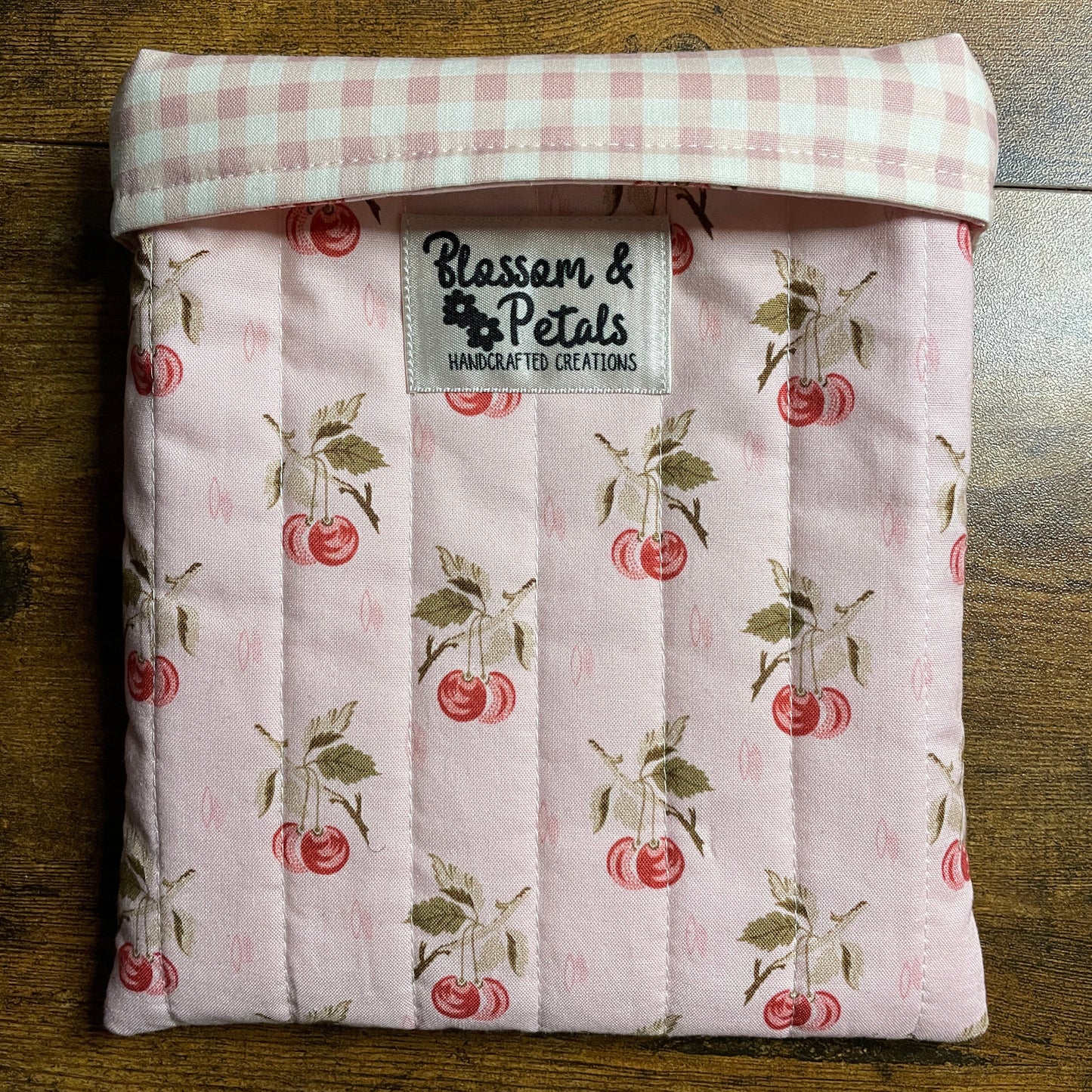 Handmade quilted pink book sleeve with cherry print fabric and Blossom & Petals label, padded fabric book cover for protecting books and journals.