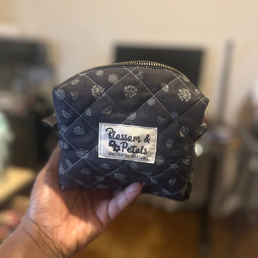 Quilted black with grey floral pouch with a visible brand label held by a hand against a blurred indoor background.